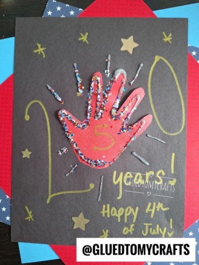 A handmade 4th of July card, perfect for 4th of July keepsakes, features a red handprint with glitter on black paper, surrounded by gold stars and "20 years! Happy 4th of July!" with @GLUEDTOMYCRAFTS at the bottom.