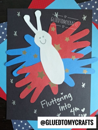 A patriotic butterfly craft made with red and blue paper handprints and a white paper body, perfect for 4th of July keepsakes. Decorated with gold star stickers on a black background, the text reads Fluttering into 4th and @GLUEDTOMYCRAFTS.
