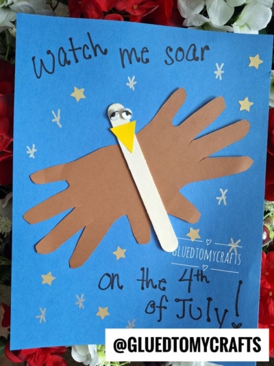 A craft showing a bald eagle made from hand-shaped brown paper and a popsicle stick on blue paper, with stars and the text Watch me soar on the 4th of July!—perfect for creating memorable 4th of July keepsakes. @GLUEDTOMYCRAFTS.