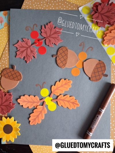 Autumn-themed paper cutouts of leaves, acorns, berries, and a sunflower are arranged on a dark gray paper with a brown marker and orange polka-dot papers in the background—perfect inspiration for FALL CRAFTS FOR KIDS. @GLUEDTOMYCRAFTS is visible twice.