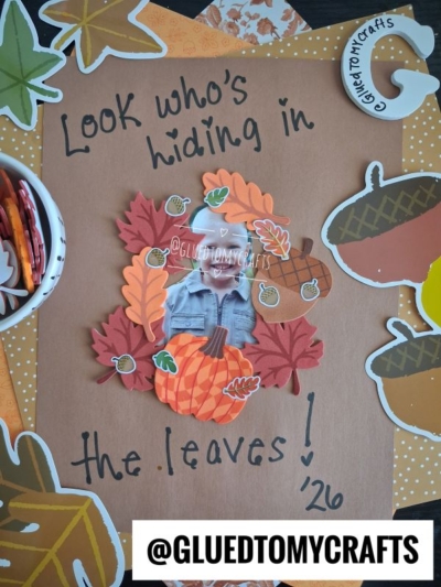 A child's photo is surrounded by paper leaves and acorns on brown paper in this charming FALL CRAFTS FOR KIDS project. The text says "Look who’s hiding in the leaves!" with “@GluedToMyCrafts” written at the bottom. Craft supplies are visible around the edges.