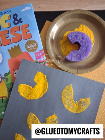 A kids’ craft scene with a yellow-painted macaroni stamp on gray paper, a gold plate with a purple sponge, a book titled “Big & Cheese,” and the handle @GLUEDTOMYCRAFTS at the bottom.