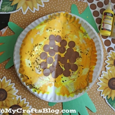 A sunflower craft made from a paper plate, yellow tissue, brown circles, and green paper handprints, with yellow and brown paint bottles nearby. Sunflower and polka dot paper decorations surround the craft.