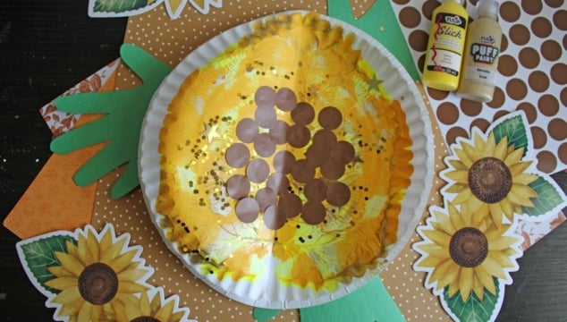 A sunflower craft made from a paper plate, yellow tissue, brown circles, and green paper handprints, with yellow and brown paint bottles nearby. Sunflower and polka dot paper decorations surround the craft.