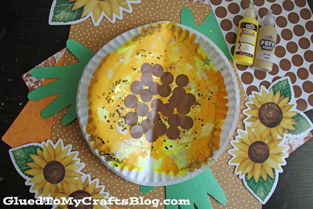 A sunflower craft made from a paper plate, yellow tissue, brown circles, and green paper handprints, with yellow and brown paint bottles nearby. Sunflower and polka dot paper decorations surround the craft.