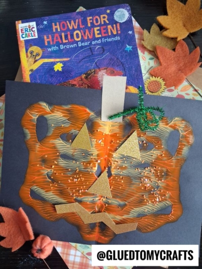 A Halloween craft featuring a tiger-shaped mask painted orange with gold triangle eyes and nose, a jagged mouth, and glitter. Perfect for halloween story time, it’s displayed on black paper with a Howl for Halloween! book and autumn decorations.