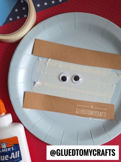A paper plate with two strips of brown paper, googly eyes, and white tape is arranged to look like a mummy—perfect for SUMMER CRAFTS FOR KIDS. Glue, tape, and craft supplies are nearby. The handle @GLUEDTOMYCRAFTS is visible.
