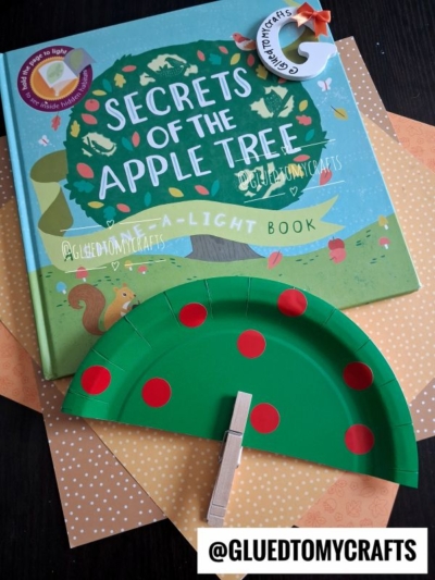 A children’s book titled Secrets of the Apple Tree, two sheets of paper, and a charming paper plate apple tree craft with red dots and a clothespin, all displayed on a dark surface.