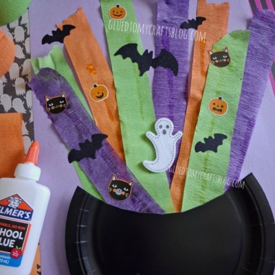Colorful Halloween-themed craft materials: strips of orange, green, and purple paper with bat, cat, and pumpkin stickers, a ghost cutout, a bottle of glue, and a black paper plate on a purple surface.