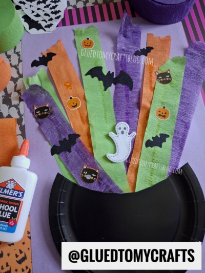 Colorful Halloween-themed craft materials: strips of orange, green, and purple paper with bat, cat, and pumpkin stickers, a ghost cutout, a bottle of glue, and a black paper plate on a purple surface.