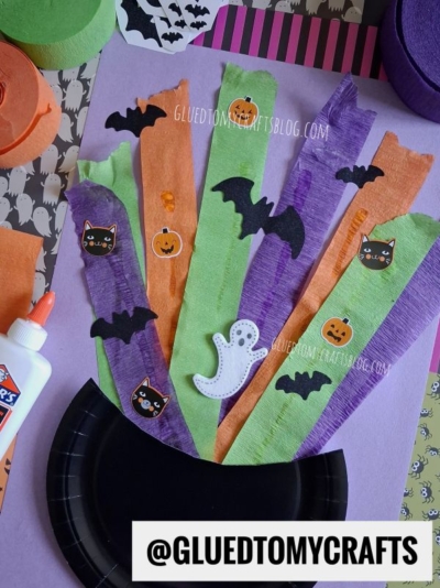 A Halloween craft with a black paper plate, orange, green, and purple streamers, and stickers of black cats, pumpkins, bats, and a ghost. Glue and craft supplies are visible around the project.