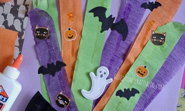 A Halloween craft with a black paper plate, orange, green, and purple streamers, and stickers of black cats, pumpkins, bats, and a ghost. Glue and craft supplies are visible around the project.