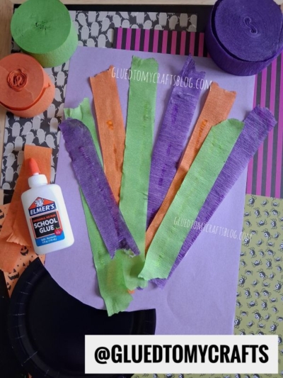 Craft supplies on a table: green, orange, and purple crepe paper strips, a bottle of Elmer’s School Glue, a black paper plate, and various patterned papers. Text reads @GLUEDTOMYCRAFTS and GLUEDTOMYCRAFTSBLOG.COM.