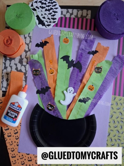 A Halloween craft scene with black plate, orange and purple crepe strips, bat, ghost, pumpkin, and cat cutouts, glue, and themed paper. The handle @GLUEDTOMYCRAFTS is displayed at the bottom.