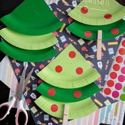 Paper plate APPLE trees with red dot stickers, clothespins for trunks, and scissors are arranged on a table with patterned paper in the background. @GLUEDTOMYCRAFTS is written on the image.