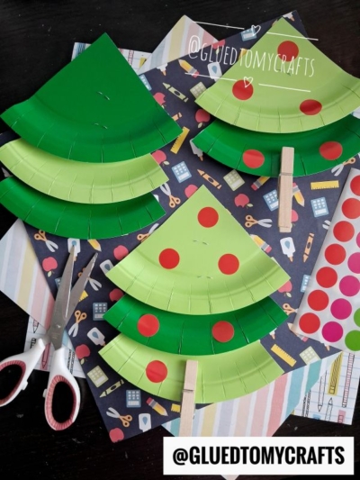 Paper plate APPLE trees with red dot stickers, clothespins for trunks, and scissors are arranged on a table with patterned paper in the background. @GLUEDTOMYCRAFTS is written on the image.