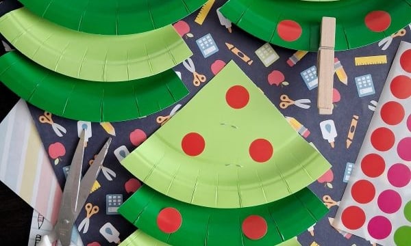 Paper plate APPLE trees with red dot stickers, clothespins for trunks, and scissors are arranged on a table with patterned paper in the background. @GLUEDTOMYCRAFTS is written on the image.