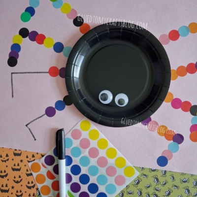A spider craft made with a black plate, googly eyes, and colorful dot stickers for legs, surrounded by Halloween-themed paper and a marker.