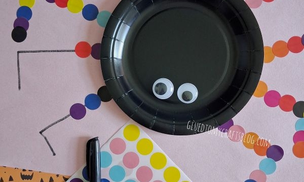 A spider craft made with a black plate, googly eyes, and colorful dot stickers for legs, surrounded by Halloween-themed paper and a marker.