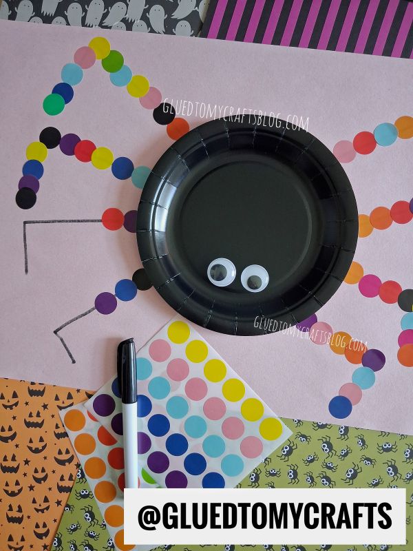 A spider craft made with a black plate, googly eyes, and colorful dot stickers for legs, surrounded by Halloween-themed paper and a marker.