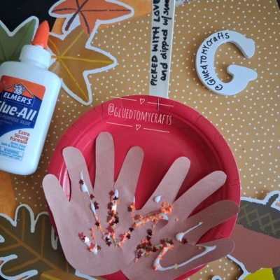 A craft setup with a red plate, brown paper hand cutouts with glued beans, a bottle of Elmer’s glue, orange star cutouts, and a popsicle stick labeled Pieced with love and dipped in sweetness—the perfect inspiration for FALL CRAFTS FOR KIDS.