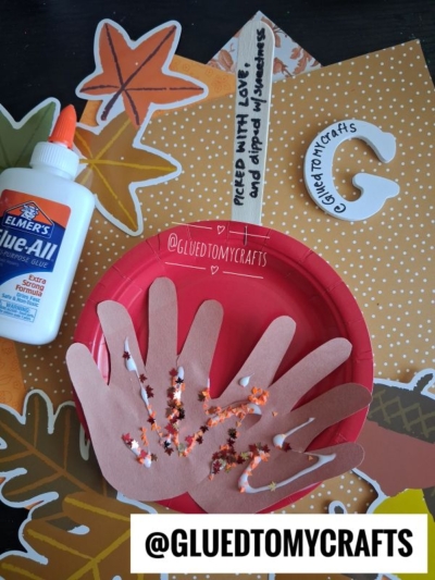 A craft setup with a red plate, brown paper hand cutouts with glued beans, a bottle of Elmer’s glue, orange star cutouts, and a popsicle stick labeled Pieced with love and dipped in sweetness—the perfect inspiration for FALL CRAFTS FOR KIDS.