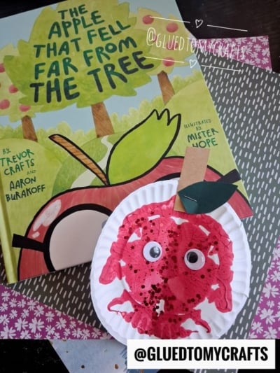 A children’s book titled “The Apple That Fell Far From The Tree” is shown with a red apple craft made from a paper plate, googly eyes, and a green paper leaf, displayed on patterned paper—perfect inspiration for PAINT SPLAT CRAFTS and other creative activities.