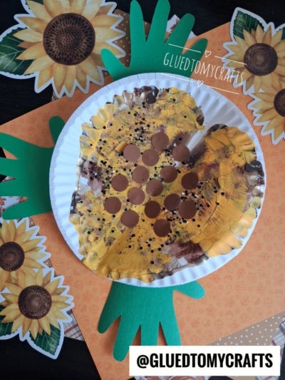 A paint splat sunflower craft made from a painted paper plate with brown dots in the center and green paper handprints for leaves, surrounded by cheerful sunflower decorations.