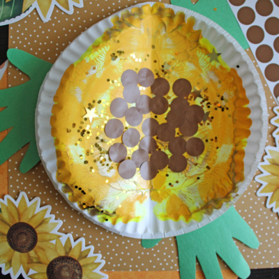A paper plate painted yellow with brown dots to resemble a paint splat sunflower, placed on top of sunflower and polka dot patterned paper, with green paper leaves attached. Text reads PAINT SPLAT SUNFLOWER.