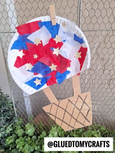 A 4th of July crafts paper plate balloon, decorated with red, white, and blue tissue paper and gold stars, is attached to a brown paper basket and hangs on a wire fence above green plants. @GLUEDTOMYCRAFTS text is at the bottom.