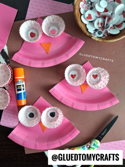 Three pink paper plate owl crafts with cupcake liner eyes and paper beaks are displayed on a table with googly eyes, a glue stick, scissors, and pink paper background. @GLUEDTOMYCRAFTS is written at the bottom.