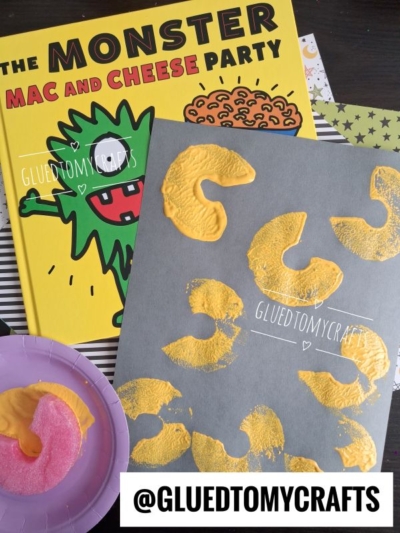 A children’s book titled The Monster Mac and Cheese Party is shown with a gray paper featuring yellow macaroni prints created using the pool noodle stamped mac and cheese technique, a pink sponge shaped like macaroni, and a plate with paint. Credit: @GLUEDTOMYCRAFTS.