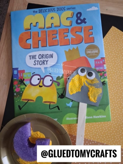 A childrens Mac & Cheese book, a gray spoon puppet with googly eyes and yellow paint, and a purple and yellow craft on a plate are on a table. The handle has @GLUEDTOMYCRAFTS written on it.