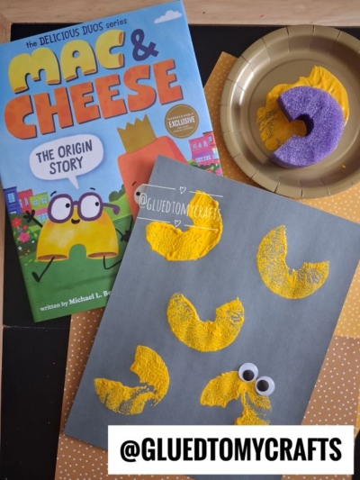 Children’s craft scene with a “Mac & Cheese” book, a gold plate holding a painted pasta noodle, and a gray paper displaying four yellow-painted macaroni shapes, two with googly eyes, on a polka-dot surface.