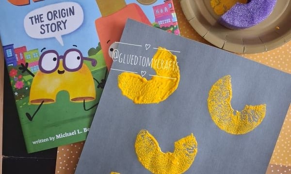 Children’s craft scene with a “Mac & Cheese” book, a gold plate holding a painted pasta noodle, and a gray paper displaying four yellow-painted macaroni shapes, two with googly eyes, on a polka-dot surface.