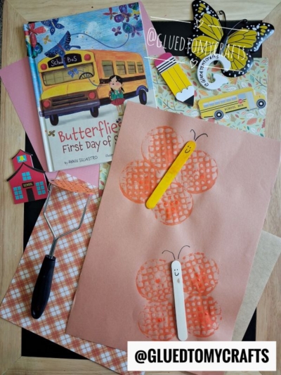 Butterfly crafts with popsicle sticks, a book titled Butterflies on the First Day of School, craft tools, and paper butterflies on a table, all arranged for a children’s art activity—including a fun potato masher stamped butterfly project.
