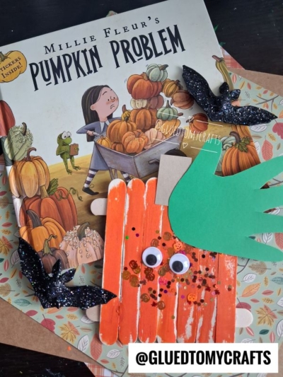 A childrens book titled Millie Fleurs Pumpkin Problem is shown with paper bats and a pumpkin craft made from orange popsicle sticks, googly eyes, green paper, and glitter—perfect for halloween story time. The @GLUEDTOMYCRAFTS handle is visible.