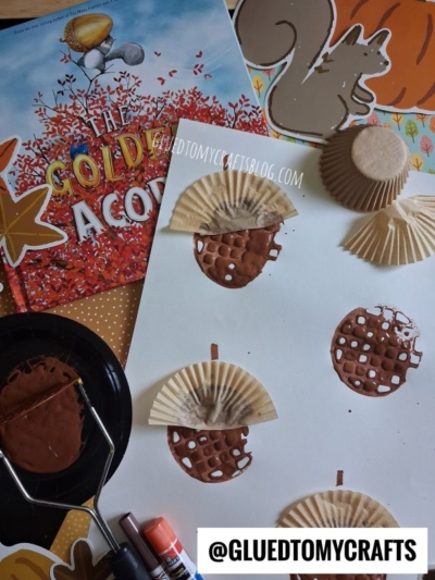Children’s acorn craft activity with a book, cupcake liners, brown paint, a painted potato masher, glue sticks, squirrel cutout, and acorn art made from cupcake liners and paint on white paper. @GLUEDTOMYCRAFTS appears at the bottom.