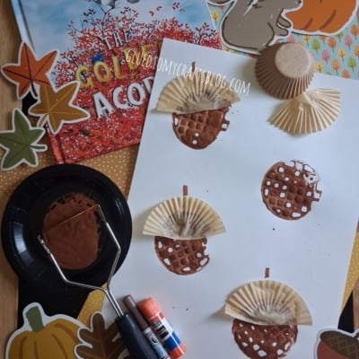 Children’s autumn craft project with painted cupcake liners as acorns on white paper, fall-themed decorations, a book titled The Golden Acorn, glue sticks, and a paint roller arranged on a table.