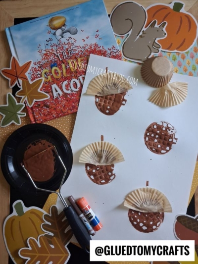 Children’s autumn craft project with painted cupcake liners as acorns on white paper, fall-themed decorations, a book titled The Golden Acorn, glue sticks, and a paint roller arranged on a table.