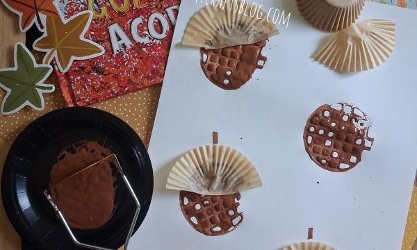 Children’s autumn craft project with painted cupcake liners as acorns on white paper, fall-themed decorations, a book titled The Golden Acorn, glue sticks, and a paint roller arranged on a table.
