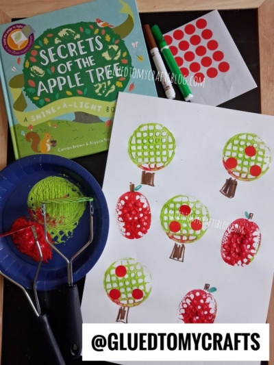 Childrens apple-themed art project featuring painted red and green apple prints—created as a delightful potato masher stamped craft—on white paper, book titled Secrets of the Apple Tree, markers, dot stickers, and painting tools arranged on a table. @GLUEDTOMYCRAFTS watermark visible.