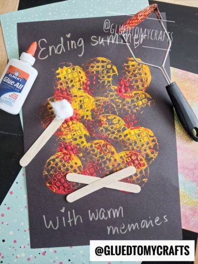 A potato masher stamped craft on black paper shows a painted campfire with yellow and red textured dots, two craft sticks as logs, a cotton ball marshmallow, and a bottle of glue. The text reads, Ending summer with warm memories.