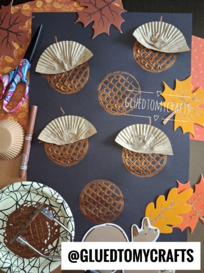 A fall craft scene with brown cupcake liners and paper circles shaped like acorns—some featuring a potato masher stamped acorn design—on black paper, surrounded by cut-out leaves, scissors, and crafting supplies. The handle @GLUEDTOMYCRAFTS is at the bottom.