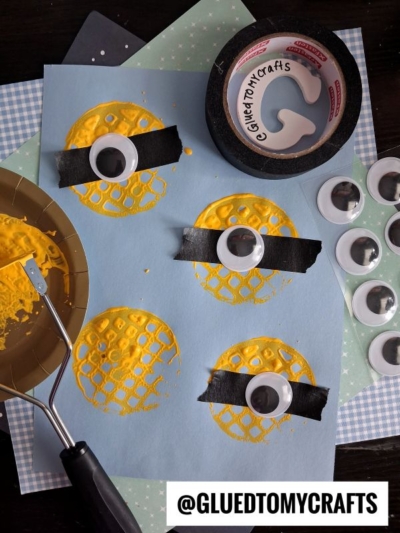 A blue paper with yellow, potato masher stamped craft circles, each featuring a large googly eye attached with black tape to resemble minions. Craft supplies like tape, paint, and extra googly eyes are nearby. Text reads @GLUEDTOMYCRAFTS.