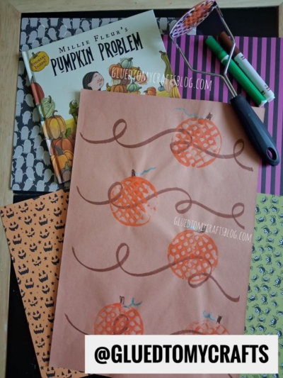 Children’s pumpkin-themed craft supplies, patterned papers, markers, a paint roller, and a book titled “Pumpkin Problem” are arranged on a table. The craft shows hand-painted pumpkins on brown paper, alongside a fun potato masher stamped craft for added texture and creativity. @GLUEDTOMYCRAFTS is at the bottom.