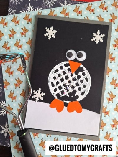 A handmade penguin card with googly eyes, orange beak and feet, and white snowflakes, made using a potato masher print on black paper, placed on festive background paper. @GLUEDTOMYCRAFTS label at bottom.