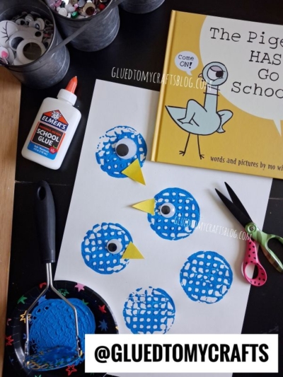 Art supplies and a children's book are on a table. Blue circle prints with googly eyes, created as part of a potato masher stamped craft, are on paper, made with glue and scissors. The book The Pigeon HAS to Go to School! is partially visible.