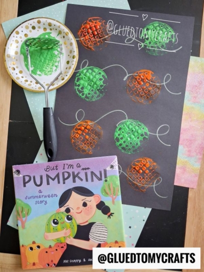 Children’s potato masher stamped craft using orange and green paint on black paper. Supplies and the book But I’m a Pumpkin! are also shown. @GLUEDTOMYCRAFTS watermark is visible.
