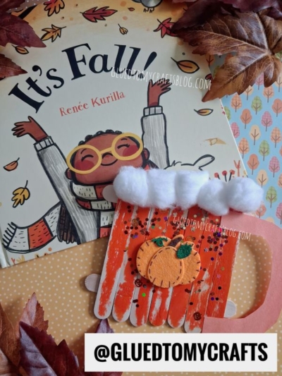 A children’s book titled Its Fall! is shown with a festive, fall-themed craft: a popsicle stick pumpkin spice mug decorated with cotton foam, a mini pumpkin, and sparkling sequins. Autumn leaves are scattered around the cozy scene.